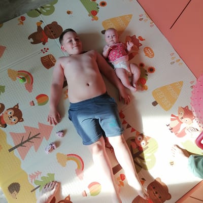 In this overhead shot, a young boy and a baby are lying on a colorful play mat patterned with cartoon forest animals and autumn leaves. The boy, who appears to be around 8-10 years old, is shirtless and wearing blue denim shorts. He is lying on his back, with his head turned to the side, looking towards the camera with a slight smile. Next to him, the baby is also lying on her back, dressed in a pink and white strawberry-patterned top and a diaper. The baby's gaze is directed upwards. The play mat is placed on a floor that appears to be wood or laminate. Sunlight streams in from the right, casting distinct shadows across the mat and highlighting the subjects. A pink, textured object, possibly a toy, is visible on the right edge of the frame. Scattered on the mat are a few small items, including tiny pink socks and what looks like a small toy rainbow with a green creature on it. The overall scene suggests a casual, indoor play session on a bright day. The setting is likely a child's room or a living area within a home. No visible text is present in the image.