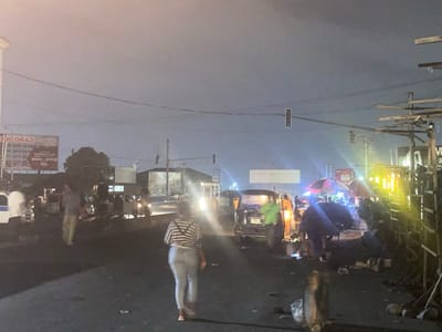 The image depicts a busy street scene at night in Port Harcourt, Nigeria. Several people are visible, including individuals walking on the road and standing near vehicles. The setting appears to be a commercial area, with a billboard and several stalls or shops lining the street. Various vehicles, including cars and what appear to be minibuses or vans, are present, suggesting ongoing traffic. The lighting is artificial, with streetlights and the headlights of vehicles illuminating the scene. Some areas are brightly lit, while others are in shadow, creating a contrast. The atmosphere appears to be active and perhaps a bit chaotic, typical of a bustling urban environment at night.
Notable details include the presence of umbrellas at some of the stalls, suggesting vendors might be operating. There are also some debris or items scattered on the ground in the foreground and mid-ground, which could indicate merchandise or refuse. The sky is dark, consistent with nighttime. There is a traffic light visible in the distance.
The billboard in the upper left corner has some visible text, including "000643" and what appears to be "BUSINESS PLACE SHOPS OFFICES". Another sign is partially visible on the right side of the image, with text including "VI" and "S". The overall impression is a lively night market or roadside commercial hub.