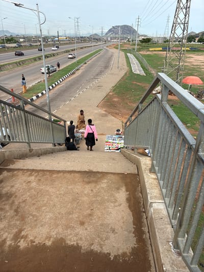 The image provides an elevated view from a pedestrian bridge overlooking a multi-lane highway and its surrounding landscape in Angwan Sayawa, Nigeria. The time of day appears to be daytime, with bright, possibly hazy or overcast conditions, as evidenced by the diffused light and lack of strong shadows.

In the immediate foreground, the galvanized metal railings of the pedestrian bridge are prominent. Directly below the bridge, on a concrete pathway, several individuals are engaged in activity. One person, wearing a pink top and black skirt, stands near a makeshift stall laden with numerous small, colorful items, suggesting a street vendor selling goods. Another person in a tan, possibly traditional, garment stands close by. Other figures are visible descending the concrete stairs of the bridge.

The highway below stretches into the distance, featuring multiple lanes separated by a grassy median with a black-and-white striped barrier. Several cars of various colors are visible on the road, with a few individuals standing on the roadside or near the median strip.

To the left of the highway, a wide expanse of grassy land is dotted with numerous tall, lattice-structured metal power transmission towers (pylons) and their connecting power lines, extending towards distant, undulating hills or mountains. A large, deflated or partially deflated orange spherical object lies in this grassy area in the mid-ground. No discernible text is visible in the image.