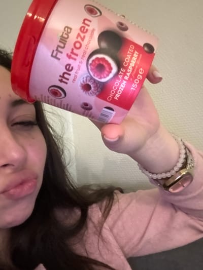 A person holds a container of "Fruita the frozen" chocolate-coated frozen raspberries. The container is pink with a red lid and features an image of the dessert and the text "real fruit & real chocolate" and "CHOCOLATE COATED FROZEN RASPBERRY". The person is wearing a pink long-sleeved shirt, a watch, and beaded bracelets. The person's face, hair, and hand holding the dessert are visible.