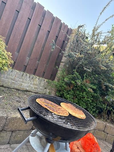A grill sits on a stone patio in a backyard in Veghel, Netherlands. Two flatbreads cook on the grill. A wooden fence and shrubs form the backdrop. An orange mesh bag rests near the grill. The sky is visible in the upper left.