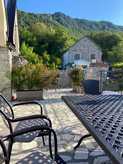 The image shows an outdoor patio in Sotonići, Montenegro. A stone house is visible in the background, nestled amongst trees and a mountain. In the foreground, there is a black metal table with a lattice design and matching chairs. A long rectangular planter with a green shrub sits along the edge of the patio. The patio itself is paved with light-colored stone. The scene is bathed in sunlight.