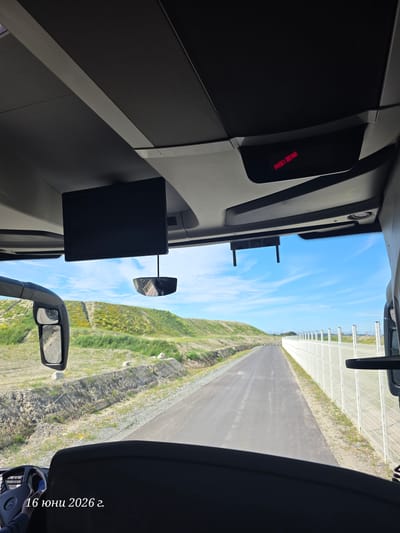 The image is taken from the interior of a vehicle, looking forward through the windshield. The view is of a paved road leading into the distance, flanked on the left by a grassy, hilly embankment with scattered rocks and on the right by a white metal fence. The sky is blue with some wispy white clouds.
In the foreground inside the vehicle, the top of a dark-colored dashboard and the steering wheel are visible on the left, with a Mercedes-Benz logo on the steering wheel. A portion of a dark seat is visible in the lower right.
Mounted on the ceiling of the vehicle, there is a black rectangular screen and below it, a small, reflective, bell-shaped object is suspended by a wire. Above the screen, a dark panel has two red rectangular indicators.
The text overlay at the bottom left of the image reads "16 юни 2026 г." which is Bulgarian for "June 16, 2026".
Based on the provided information, the location is identified as Le Havre, France. This suggests the image was captured in an industrial or construction area within or near the city, characterized by the road, embankment, and fencing, possibly related to port expansion or development. The date indicates a future capture.