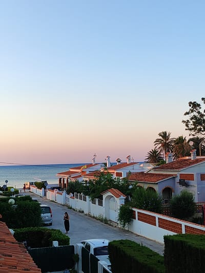 The image depicts a street scene in Els Poblets, Spain, at sunset. The sky transitions from a pale blue at the top to soft pink and orange hues near the horizon, where the Mediterranean Sea is visible. A row of white houses with terracotta tile roofs lines the street, many featuring arched windows and doorways. Lush green vegetation and palm trees are interspersed throughout the properties. On the street, a woman in a black dress stands near a silver minivan. Further down the street, another vehicle and a pedestrian can be seen. The overall atmosphere suggests a peaceful, residential coastal town.