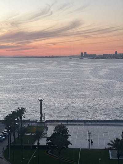 A scenic view of the Kordon coastline in İzmir, Turkey, taken at dawn or dusk, is captured from an elevated perspective. A colorful, multi-hued sky transitioning from white to orange stretches across the left side of the frame, indicating either sunrise or sunset. The Aegean Sea occupies the central section of the image, its surface reflecting the sky. A city skyline is visible along the distant shore, with building silhouettes suggesting a dense urban area.

The right side of the image shows the Kordon’s promenade. A pedestrian walkway runs along the waterfront, with palm trees interspersed. A statue or monument is located near the waterfront. A paved area extends inland from the promenade, with people and bicycles visible. Adjacent to the paved area is a strip of green space, likely a park or lawn, with more trees and landscaping elements. Cars are parked in the lower right corner.