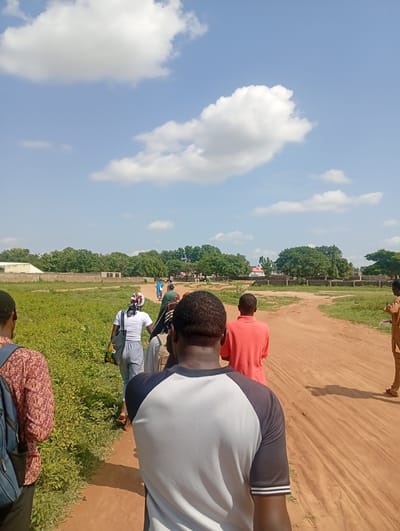 This image depicts a group of people walking on a dirt path in a rural or semi-urban area, likely in Nassarawa, Nigeria. The environment is characterized by dry, reddish-brown earth forming the path, with green, overgrown vegetation on the left side of the frame. In the background, there are low-lying buildings, some with concrete walls and roofs, interspersed with lush green trees and a wall. The sky is predominantly blue with scattered white clouds.
Several individuals are visible, mostly from behind, walking in the same direction along the path. In the foreground, the back of a person wearing a white and grey t-shirt is prominent. To their left, a person in a patterned shirt with a blue backpack is partially visible. Ahead on the path, a person in a white t-shirt and grey pants is walking, carrying a bag. Further ahead, a person in an orange t-shirt and another in a green shirt are visible. Towards the right side of the image, a person in a brown outfit is standing. The people appear to be moving as a group, suggesting a shared activity or journey. The overall impression is one of movement through an open, somewhat undeveloped area under a bright, sunny sky.