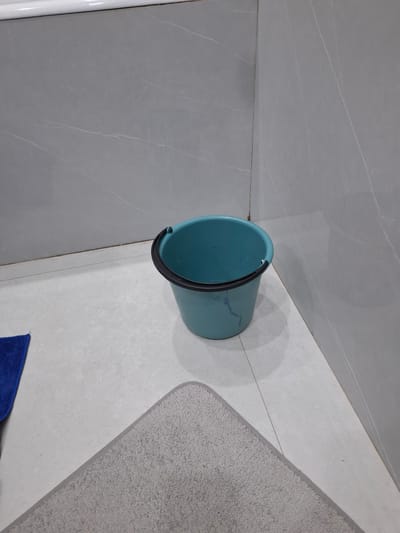 A teal bucket with a black rim sits on a light gray tiled floor in a bathroom corner. The walls are covered in large, gray marble-patterned tiles. To the left of the bucket, a corner of a blue microfiber cloth is visible. A light gray bath mat with a darker gray border is positioned in the foreground, partially obscuring the floor. The scene appears to be indoors in a residential setting, likely a bathroom, in Kasakh, Armenia. There are no people or animals present. The lighting is neutral, suggesting an indoor light source or diffused natural light.