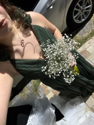 A person lies down, wearing a green pleated dress and a gold necklace with intertwined circles. They hold a bouquet of baby's breath flowers in their hands. Next to them is a wedding dress with white lace and black accents, and a car tire visible in the background. The ground appears to be a mix of pavement, grass, and other debris.
