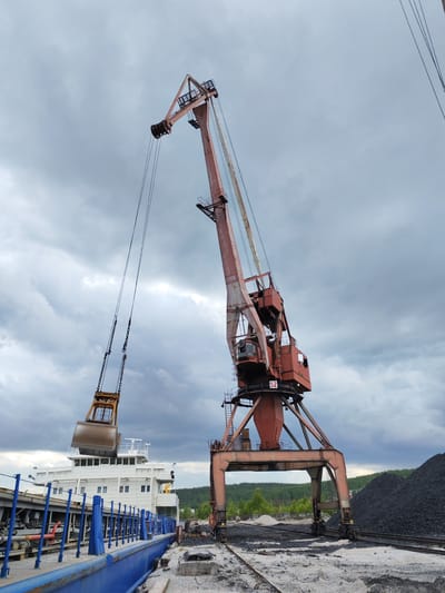 The image depicts an active industrial port scene in Medvezhyegorsk, Russia, under a heavily overcast sky. The primary subject is a large, reddish-orange gantry crane, exhibiting signs of wear and heavy use, positioned centrally. Its imposing structure features multiple levels and operator cabins, with the number "52" visible on its lower section.

Suspended from the crane's cables is a heavy-duty excavator bucket or grabber, colored brown/gold. This grabber is positioned directly above the deck of a large, white ship or vessel, indicating ongoing or imminent bulk cargo handling operations, such as loading or unloading. The ship, with its multi-story white superstructure and blue deck railings, is docked alongside a pier.

On the ground to the right of the crane, large piles of black, granular material, consistent with coal or other bulk commodities, are visible. Multiple rail tracks run beneath the crane's gantry base and along the waterfront, suggesting railway transport integration within the port's logistics. In the distance, a dense green forest covers rolling hills, contrasting with the industrial foreground.

The weather is characterized by a thick, gray, cloudy sky, suggesting an overcast daytime. No individuals are visible in the frame. The scene conveys a sense of large-scale industrial activity and transportation of bulk goods in a northern, waterfront setting.