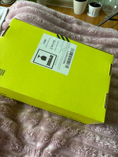 An unboxing scene is captured in this top-down, medium shot image, featuring a bright yellow cardboard box partially obscuring a soft, pink, fuzzy blanket. The box is angled towards the viewer from the left side of the frame. A white label with black text and a barcode is affixed to the top of the box. Text on the label indicates the location as "Novoe Devyatkino, Russia" and includes details like "Courier Service," "Order Number," and delivery information. A graphic of a wine glass within a square frame is also present on the label, suggesting the contents are fragile. The background shows a wooden floor with several potted plants.