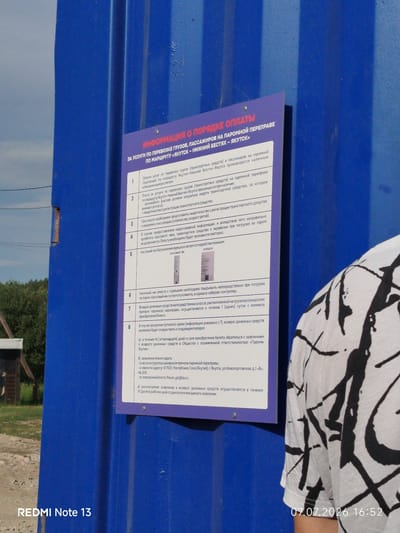 A person wearing a white t-shirt with black abstract paint strokes stands in front of a blue metal wall. On the wall, a purple sign displays information about ferry payment procedures for passenger and cargo transportation on the route "Yakutsk - Nizhny Bestyakh - Yakutsk." The sign is written in Russian. The bottom left corner of the image shows "REDMI Note 13," and the bottom right corner displays the date "07.07.2026 16:52." The background consists of greenery and a wooden structure, suggesting an outdoor setting in a place like Nizhny Bestyakh, Russia, likely near a ferry terminal.