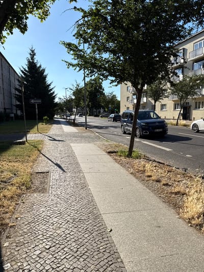 This image depicts a street scene in Rathenow, Germany, on a clear day. The perspective is from a sidewalk looking towards the street and adjacent buildings.

In the foreground, a cobblestone sidewalk runs along the right side of the image, bordered by a strip of dry grass. Beyond the sidewalk, a paved street is visible, with several cars parked along its edge and in a lane of traffic. The cars are of various colors, including dark blue and white. The street is lined with trees, some of which are full of green leaves, casting shadows on the pavement.

On the left side of the street, a multi-story building with apartment windows and balconies is visible. The building appears to be modern in design. Further down the street, more trees and buildings can be seen, suggesting an urban or suburban environment.

A blue directional sign with white text and an arrow is present on the left side of the street, indicating a street name. Traffic lights are also visible in the distance, along with streetlights.

The time of day appears to be daytime, likely morning or afternoon, given the bright sunlight and distinct shadows. The weather seems to be fair and sunny. There are no people visible in the image. The scene conveys a sense of everyday urban life in a European city. The license plate on the dark blue car reads "HVL-WO 725".