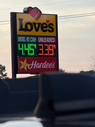 This is a close-up shot of a Love's gas station sign and part of a Hardee's sign below it. The Love's sign is yellow with black lettering that says "Love's" in large font, and a red heart logo above it. Below the "Love's" text, there are two rows of digital numbers. The first row shows "DIESEL #2 CASH" in green text and "UNLEADED" in red text. The second row shows "4.45" in green digits and "3.39" in red digits, indicating gas prices. The Hardee's sign is red and displays the Hardee's logo of a yellow star with a smiling face, and the text "Hardee's" in white script. The image is taken at eye level, looking slightly upwards, with the gas station sign in focus. The background shows a pale sky, some power lines, and the tops of trees. The lower part of the image is blurred and dark, suggesting it is a view from inside a vehicle. The location is identified as Piedmont, United States of America.
