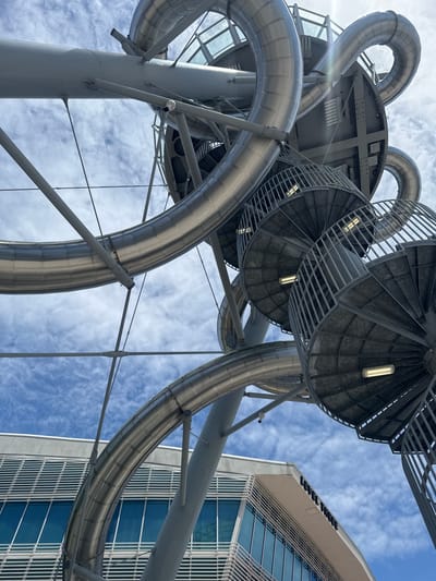 A low-angle outdoor photograph captures a large, modern, metallic spiral slide structure against a cloudy blue sky. The slide is made of polished metal tubes that curve and twist in complex formations, supported by a grey steel framework. Multiple circular, metal spiral staircases with open railings are visible, integrated into the structure, with industrial lights illuminating the steps.
Below the slide structure, the upper section of a contemporary building is visible. This building features a light-colored facade with prominent horizontal louvers and large, blue-tinted glass windows. Text is visible on the building's exterior, reading "LEVEL THREE". The scene appears to be in the city of Aventura, United States of America. No people are present in the frame.