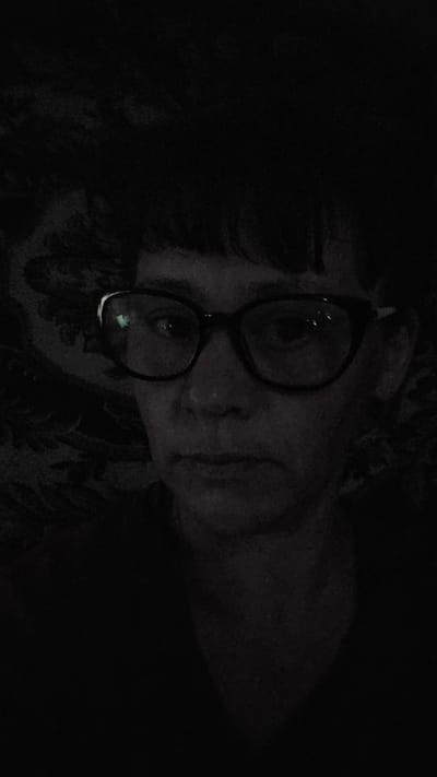 The image shows a close-up, dimly lit selfie of a person wearing dark-rimmed glasses. The person's face is visible from the nose up, with their mouth slightly open. The background is dark and appears to have a textured pattern. There is a reflection in the glasses, suggesting artificial light. The location is identified as Vozsiiatske, Ukraine.