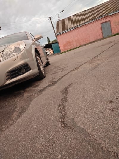 A silver car is parked on an asphalt road in Rzhyshchiv, Ukraine. The car is in the foreground, angled towards the viewer, with its front left wheel and bumper clearly visible. Behind the car, a pink brick building with a corrugated metal roof and two gray doors can be seen. Utility poles and power lines are visible against a cloudy sky. The road surface is rough and shows signs of wear, with some dark patches that may indicate water or oil stains. The overall scene suggests an outdoor, urban or semi-urban environment.