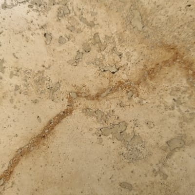 The image displays a close-up view of a textured, light brown, and beige surface, possibly concrete or aged plaster. Irregular darker patches and a reddish-brown linear stain are visible, suggesting wear, damage, or water staining. The surface has a rough, granular texture with small pores and embedded lighter-colored fragments. There are no people, objects, or discernible actions in the frame. The context of Jerez, Spain, is not visually represented in this abstract detail shot of a surface.