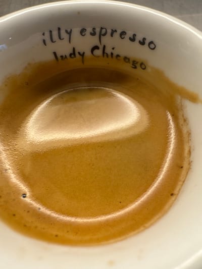 The image displays an overhead view of a white ceramic espresso cup, located in Andratx, Spain. The cup contains espresso, characterized by a rich, reddish-brown crema surface that reflects an overhead light source. Inscribed in black script on the interior rim of the cup are the words "illy espresso" on the upper line and "Judy Chicago" on the line below. No individuals, specific actions, or interactions are visible within the frame.