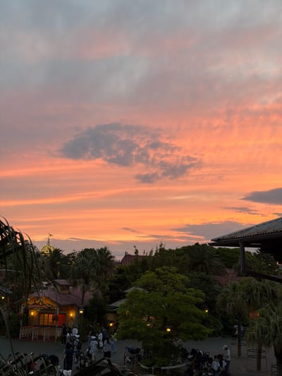 The image captures a sunset scene in Tokyo Disneyland, Urayasu, Japan. The sky is filled with vibrant orange and pink hues, with scattered grey clouds. In the foreground, lush green foliage partially obscures the view. In the mid-ground, several buildings with rustic architecture are visible, including a prominent structure with a red roof and illuminated windows. A dome-shaped building with a yellow flag on top is also noticeable. A group of people, some with strollers, are walking around a paved area in front of the buildings. The overall atmosphere suggests a theme park setting during dusk.