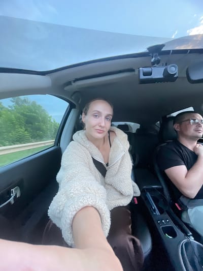 The image shows two people in the front seats of a car, likely traveling on a road in Pirogovo, Russia. The woman in the foreground is taking a selfie, wearing a fluffy cream-colored jacket over a black top. She has blonde hair tied back and is looking at the camera with a slight smile. To her right, in the passenger seat, a man wearing glasses and a black t-shirt is visible. He is looking forward, seemingly driving or in the passenger seat. The car's interior is visible, including the dashboard and a sunroof that lets in the daylight. Outside the car, lush green trees line the road, suggesting a rural or suburban setting. A dashboard camera is mounted near the sunroof.