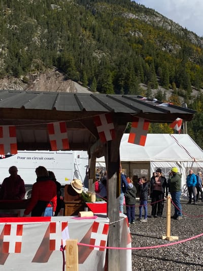 The image depicts an outdoor market scene in Abondance, France, situated at the base of a forested mountain. In the foreground, a wooden market stall with a dark corrugated roof is prominently featured. Strings of red flags with white crosses, resembling Swiss flags, are strung beneath the stall's roof and along its front counter. A pink braided rope barrier, secured by wooden posts, delineates the immediate stall area in the lower right.
Behind the stall counter on the left, three adults are present; one wears a dark red jacket, and another, facing away, wears a red jacket. Behind them, a white truck is visible with the French text "Pour un circuit court." printed on its side. On the stall's counter, a straw hat, several glass jars, and what appears to be a block of cheese are displayed.
To the right of the stall, a group of approximately ten individuals, including adults and children, stand on a gravel surface. Notable figures include two children in dark jackets, a woman with folded arms, and a man wearing a green jacket and a yellow beanie. In the midground, a large white marquee tent is erected to the right of the wooden stall, accommodating more people further back. The background is dominated by a steep, tree-covered mountainside composed primarily of conifers, with visible patches of exposed rock and soil. The sky above is partly cloudy.