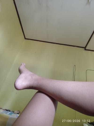 The image shows a close-up view of a person's legs and feet. The legs are crossed, with the top leg resting over the bottom leg. The skin appears smooth and has a light tan complexion. The background consists of a plain yellow wall and a white ceiling. A date and time stamp in the lower right corner reads "27/06/2026 13:12". There are no visible objects or people other than the legs. The image does not provide enough information to determine the specific location within Sukaraja, Indonesia.