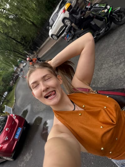 A young woman with blonde hair pulled into a ponytail is taking a selfie outdoors. She is wearing an orange halter top adorned with gold studs and has her right arm raised, with her hand in her hair. Her eyes are closed, and her tongue is sticking out. She is standing on a wet asphalt road with puddles reflecting the overcast sky. In the background, a red car with a blue license plate reading "A 4A22J" is parked on the left side of the road. Further down the road, a white SUV is driving, and behind it, a person in black riding a green motorcycle is visible. Trees line the left side of the road, and a black metal fence runs parallel to the street. The scene appears to be a street in Xiangfang District, China, on a rainy day.