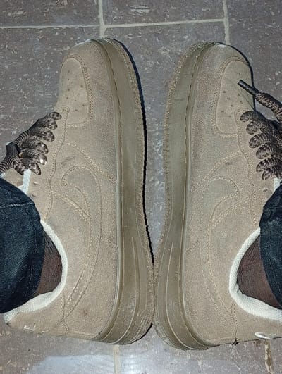 The image displays a pair of dusty, tan suede Nike Air Force 1 shoes. The shoes are positioned side-by-side, facing inwards, on a tiled floor with a speckled gray pattern. The laces are dark brown and patterned. The shoes appear worn, with visible scuff marks and dirt accumulation, particularly on the soles and the suede upper. Dark denim trousers are visible around the ankles of the wearer. The scene is likely indoors, given the tiled flooring. The context of Ukpor, Nigeria, suggests this might be a common type of footwear for the local environment.
