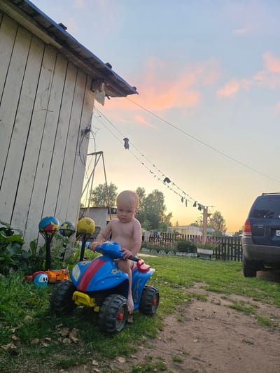 A young, shirtless boy with blonde hair sits on a blue and red toy ATV in a grassy area next to a white wooden building. The boy is barefoot and facing forward, with his hands on the handlebars. In the background, there is a fence with flowers, trees, a dark SUV, and a clothesline with items hanging on it. The sky is a pale orange and pink, indicating sunset. The setting appears to be a rural area, possibly the city of Буренка, Russia.