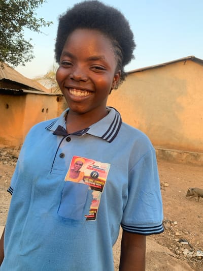 A smiling girl stands outdoors, wearing a light blue polo shirt with dark blue trim. Patches, one featuring a man's portrait and the text "2 Years Remembrance" are affixed to her shirt. A building is partially visible in the background, and a primate can be seen at the top of the image. The photo appears to have been taken during daylight hours, with natural light illuminating the scene.