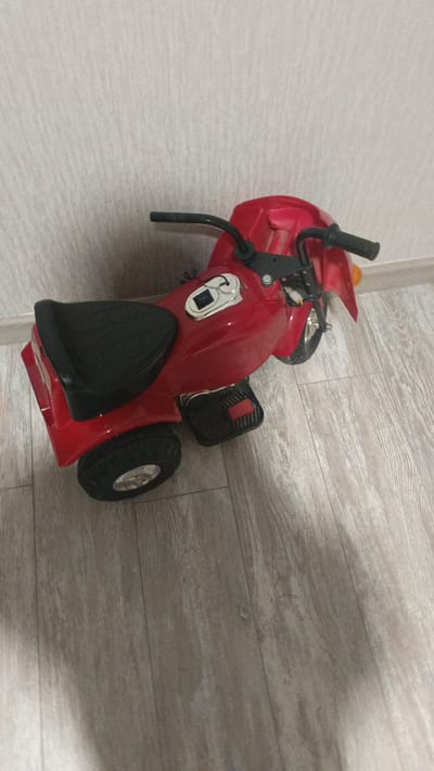 A red toy motorcycle is on a light-colored wooden floor against a textured, light-colored wall. The motorcycle has a black seat, black handlebars, black footrests, and two black wheels. A silver switch is visible on the top of the motorcycle. The scene appears to be indoors, possibly in a residential setting in Aksinyino, Russia.
