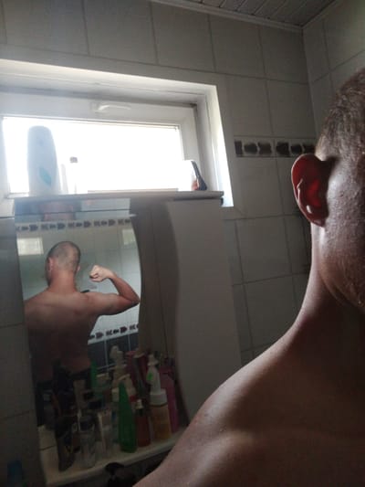 The image depicts a bathroom interior, possibly in Kirensk, Russia. The foreground is dominated by the shoulder and ear of a person, suggesting a selfie taken in a mirror. In the background, a mirror reflects a man's back, with his left arm flexed to display his bicep. The bathroom features tiled walls and a window above a shelf cluttered with various bottles and toiletries. The lighting appears to be natural, coming from the window.