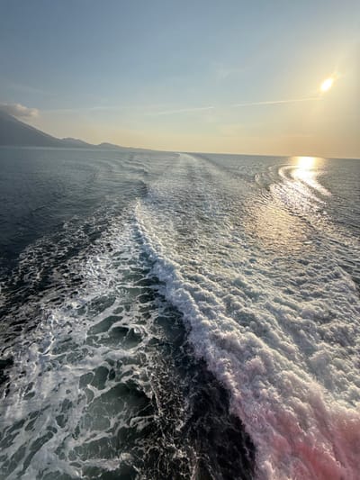 The scene depicts the turbulent wake of a moving vessel at sea near Palaiopoli, Greece. The foreground and midground are occupied by dark blue-green water agitated into white foam and waves by the boat's propulsion, with a notable reddish tint visible within the foam on the right side of the wake. A distinct horizon line separates the water from the sky. In the background, a range of mountains or landmasses is visible on the left, appearing dark and hazy against the sky. The sun is positioned low in the upper right quadrant of the sky, casting a bright glare and creating a prominent path of reflection across the water surface towards the viewer. The sky above is clear, transitioning from pale blue overhead to a golden hue near the horizon where the sun is located. No individuals or other discernible objects are present.