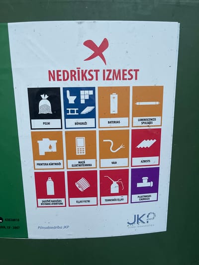 The image shows a poster with various icons and text in Latvian, indicating items that should not be discarded. The poster is mounted on a green surface, likely a waste bin or a wall. The text at the top reads "NEDRĪKST IZMEST," which translates to "DO NOT DISCARD." Below this are several squares with icons and labels, representing different categories of waste. These include:
- A bag of trash labeled "PELNI" (Ashes)
- Icons related to construction materials labeled "BŪVGRŪŽI" (Construction Debris)
- A battery labeled "BATERIJAS" (Batteries)
- A fluorescent light bulb labeled "LUMINISCENCES SPULDZES" (Fluorescent Bulbs)
- A printer cartridge labeled "PRINTERA KARTRIĜI" (Printer Cartridges)
- Small electronics labeled "MAZĀ ELEKTROTEHNIKA" (Small Electronics)
- A coiled wire labeled "VADI" (Wires)
- Asbestos labeled "AZBESTS" (Asbestos)
- A spray can labeled "ĢĀDZĪVĘ RADUŠIES BĪSTAMIE ATKRITUMI" (Damaged Hazardous Waste)
- Oil filters labeled "EĻĻAS FILTRI" (Oil Filters)
- Technical oils labeled "TEHNISKĀS EĻĻAS" (Technical Oils)
- Plastic pipes labeled "PLASTMASAS CAURULES" (Plastic Pipes)
In the bottom left corner, there is contact information: "63026010" and "AVA, LV - 3007." This suggests the location is in the city of Jelgava, Latvia, as "LV" denotes Latvia and "AVA" is likely an abbreviation or street name within Jelgava, and "3007" is a postal code. The text "Pilnsabiedrība JKP" and a logo with "VIDEI DRAUDZĪGS" (Environmentally Friendly) are also present. The overall scene is a close-up of a waste disposal information poster.