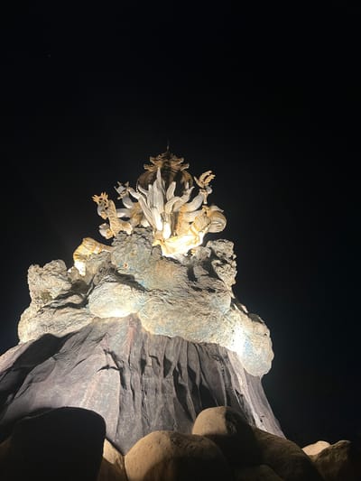 In Kerobokan Kelod, Indonesia, a large, intricately carved monument stands illuminated against a dark night sky. The base of the monument consists of rugged, dark gray and brown rock-like formations, resembling a stylized mountain or cliff. The upper section features multiple light-colored, sculpted figures, likely deities, with prominent gold ornamentation and crowns. These figures are dynamically posed, some exhibiting multiple arms, and are brightly illuminated from below with a warm, yellowish light. A complex golden structure crowns the uppermost part of the sculpture. The intense illumination creates stark contrasts with the surrounding deep black sky. No individuals are present, and no actions or interactions are visible beyond the static representation of the sculpture.