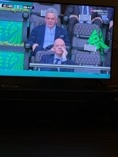 The image is a television screen displaying a live broadcast of a football match. In the foreground, two men are seated in plush chairs. The man on the left, with curly gray hair, wears a dark blue suit and a light blue shirt. The man on the right, with a bald head, wears a dark suit and a blue tie. Both men appear to be officials or dignitaries, with nameplates visible on the chairs. A green flag with intricate cutouts, bearing the FIFA logo, is placed on the chair of the man on the right. The background shows a stadium with green decorative elements and a score display at the top left corner indicating "MEX 1 0 RSA". The "FIFA WM 2026 GRUPPE A" text is visible on the upper right side of the screen. The overall scene is a sports event, likely a FIFA World Cup match. There is no indication of a location in the city of Hüsen, Switzerland.
