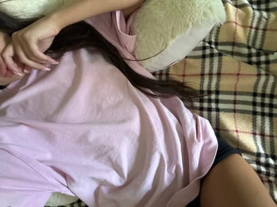 An overhead shot shows a person in bed. The person is lying on their side, facing away from the camera, with their face partially obscured by their hand. They are wearing a pink t-shirt and black shorts, and their dark hair is spread across the pillow and t-shirt. The person is lying on a plaid blanket. A fluffy white pillow is behind their head. The scene appears to be indoors, likely in a bedroom. The location in Basarabeasca, Moldova is not discernible from the image.