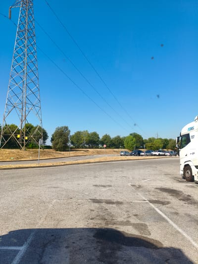 A photograph taken on a sunny day shows a parking lot in Chiari, Italy, with a large metal pylon in the background and a white truck in the foreground. The pylon, a lattice structure of metal beams, stands tall against a clear blue sky, with several power lines extending from it. The parking lot is paved with asphalt and marked with white lines, indicating parking spaces. In the middle ground, a line of cars is parked. The environment is mostly dry, with some patches of grass and trees visible. The overall scene depicts an outdoor commercial or industrial area.