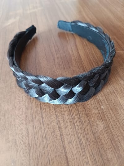 A close-up shot shows a black braided headband with faux hair on a wooden surface. The headband is curved, with the braided portion prominently displayed in the foreground. The wood grain of the surface is visible, running diagonally from the bottom left to the top right of the image. There is no indication of location within the city of Tebras, Latvia. The scene is static and shows no people or actions.