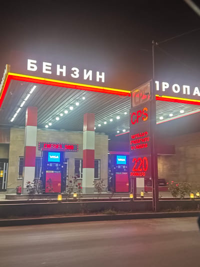 This is a night-time, outdoor photograph of a gas station in Armavir, Armenia. The scene is illuminated by the bright neon lights of the station's signage. The primary subjects are the gas pumps and the overhead signage displaying fuel prices and types.

The vertical signage features large, white, illuminated letters spelling out "БЕНЗИН" (Benzin, Russian for Gasoline) on the left side. To the right, a tall pylon displays digital prices. The top section shows "CPS" in red letters, followed by what appears to be Armenian script in red text. Below this, a prominent red digital display shows "220" with "ТРАКТИР" (Traktir, Russian for Tavern/Inn, possibly indicating a fuel type or promotion) in smaller red text underneath. Further down, a screen displays "VISA" in blue and white.

Another, lower sign displays "DIESEL 560" in red digital letters, with a "VISA" logo below it. The gas pumps themselves are red and white. The overall setting appears to be a typical modern gas station, with a covered canopy over the pumps.

The image is taken at night, as indicated by the darkness of the sky and the reliance on artificial illumination. There are no people visible in the photograph. The weather appears clear, with no visible rain or fog. The presence of Armenian script and the mention of Armavir place the location specifically. The overall mood is functional and commercial, highlighted by the bright, attention-grabbing lights of the fuel prices.