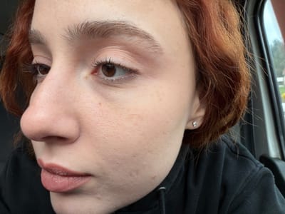 A close-up profile view shows a young adult female inside a vehicle in Plaine, France. The individual has fair skin with light freckles, reddish-brown hair, and brown eyes with visible eyelashes and light-colored eyeshadow. Her eyebrows are light brown and full. A small, clear stud earring is visible in her right earlobe. Her lips are a light pink-mauve shade. She is wearing a dark-colored hooded garment. The background through the vehicle window displays blurred green foliage and indistinct structures, suggesting an outdoor environment.