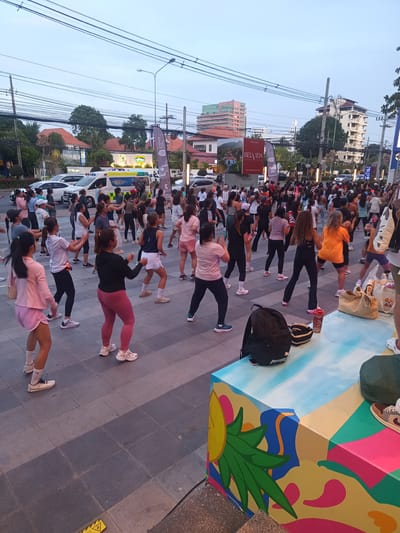 A daytime scene in Hua Hin City Municipality, Thailand shows a large group of people participating in an outdoor exercise class on a paved plaza. An ambulance is parked on the street behind the crowd. Buildings are visible in the background, including a high-rise structure bearing the name "BELVIDA." A painted, cubic structure is in the foreground, with a backpack and other personal items resting on top. The participants, mostly women, are dressed in workout attire, and are engaged in synchronized movements. Overhead power lines are visible in the sky.