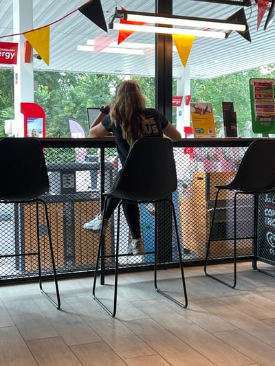 In Dilbeek, Belgium, at a gas station with an attached convenience store and seating area, a young woman with long, wavy brown hair is seated at a counter, facing away from the camera. She is wearing a black t-shirt with white lettering and black leggings, along with light-colored sneakers. A laptop is open in front of her on the counter. The seating area features two black bar stools, one of which is empty and to the left of the woman. The counter is behind a black metal mesh partition, and beyond it, large windows look out onto a treed area. Overhead, fluorescent lighting fixtures and colorful triangular flags are visible. Signs for "Energy" are visible on a white pillar to the left. The floor is made of light-colored wood-look tiles.
