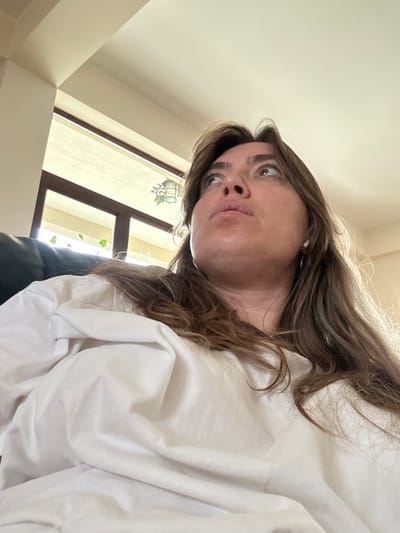 A low-angle, medium close-up shot depicts a female subject positioned indoors in the city of Sighnaghi, Georgia. She possesses shoulder-length, light brown hair with lighter streaks and wears a loose white garment. A white earbud is visible in her right ear. Her head is tilted slightly back, and her gaze is directed upwards and to her right, with a neutral expression. The background features a bright, light-colored ceiling on the upper left. To her left and behind her, a large window or glass door with dark frames allows ambient light into the space. Visible through the window are indistinct green plants, one appearing to be in a hanging basket, and a cream-colored roller blind.