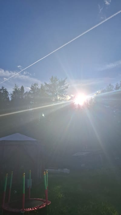 The image depicts a backyard scene on a clear, sunny day in Novovilgovskoye Sel'skoye Poseleniye, Russia. The sun is low in the sky, casting bright rays and creating lens flare across the frame, partially obscuring the background. A contrail from an aircraft is visible stretching diagonally across the bright blue sky. In the foreground, a red and green trampoline is set up on a grassy lawn. To the left of the trampoline is a light-colored gazebo. Behind the gazebo, a dense line of trees forms a dark silhouette against the bright sky. Further back, a small structure, possibly a shed or outbuilding, is visible among the trees. The overall impression is that of a tranquil outdoor setting.