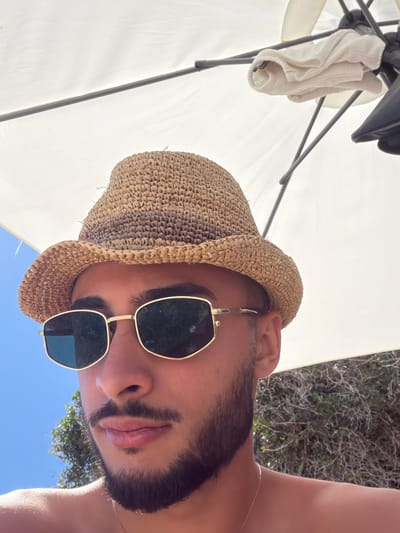 A close-up, medium shot captures a young man wearing a straw fedora and gold-rimmed sunglasses. He is looking downwards and slightly to his right. The man has a beard and mustache. He is shirtless, and a thin gold chain is visible around his neck. He is positioned outdoors, under what appears to be a large white umbrella, with a clear blue sky visible in the background, along with some foliage and greenery. The overall setting suggests a sunny day, possibly at a beach or outdoor recreational area within Elaties Municipal Unit, Greece.