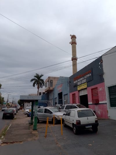The image depicts a street scene in Cuiabá, Brazil, under an overcast sky. The foreground shows a paved road with a sidewalk and a bus stop shelter. Several cars are parked along the street, including a dark-colored sedan, a silver SUV, and a white hatchback.

Dominating the background is a commercial building with a facade painted in shades of gray, pink, and blue. A prominent sign on the building reads "KOYAMA AUTO CENTER INJEÇÃO, MECÂNICA, ELÉTRICA 3623-6380". Below this, another sign advertises "FAROL, TROCA DE ÓLEO, CÂMBIO AUTOMÁTICO". The building appears to be an auto repair shop.

Further in the background, a tall industrial chimney with multiple antennas or antennae protrudes into the sky. A palm tree is visible to the left of the chimney, situated between buildings. Utility wires are strung across the scene, contributing to the urban landscape. No people are clearly visible in the image.