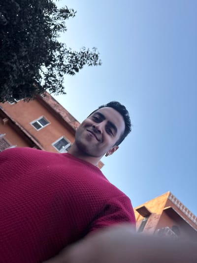 A low-angle photograph depicts a young adult male positioned in the lower central frame, looking directly towards the camera with a subtle smile. He has dark hair and wears a dark red, waffle-knit long-sleeved garment. A metallic earring is visible in his left ear. Blurred fingers are present in the lower foreground, indicating proximity to the camera lens. The background features two terracotta-colored buildings, consistent with the architectural style found in Jaipur, India. The structure on the left is multi-storied with two visible windows, partially obscured by dark green tree foliage in the upper left. A second building on the right exhibits a decorative white roofline frieze, silhouetted against a clear, light blue sky. The scene is illuminated by natural daylight.
