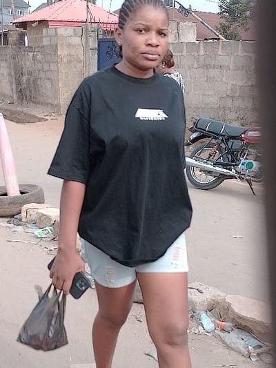 A woman wearing a black shirt and denim shorts stands outdoors. She holds a phone and a black plastic bag in her hand. Behind her is a parked motorcycle and a brick building. The ground appears to be dirt or unpaved. The image is slightly rotated, causing a skewed perspective.