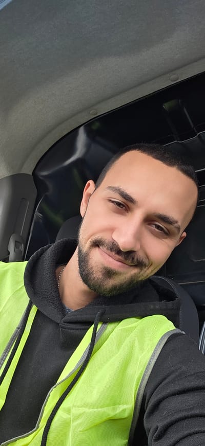A man is posing for a selfie. He is wearing a black hooded sweatshirt, a lime green reflective safety vest, and a silver chain around his neck. He has short dark hair, brown eyes, and a trimmed beard. He is smiling slightly at the camera. The interior of a vehicle is visible behind him. The location is unknown and the city of Crețuleasca, Romania, cannot be verified from the visual information provided.